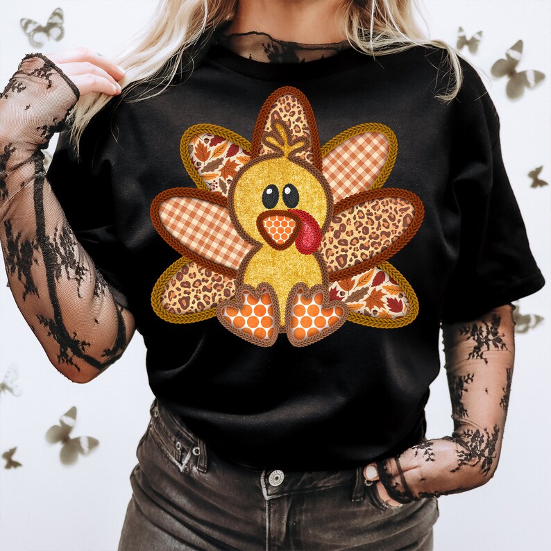 May include: A black t-shirt featuring a cute cartoon turkey design. The turkey is made from various patterned fabrics, including plaid, leopard print, and leaves. The turkey is wearing a red wattle and has a yellow body.