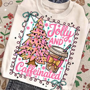 Puede incluir: Sudadera color crema con un diseño de árbol de Navidad rosa, adornado con luces de colores y manchas negras. El texto "Jolly AND Caffeinated" se muestra con un gráfico de taza de café. El diseño está enmarcado.