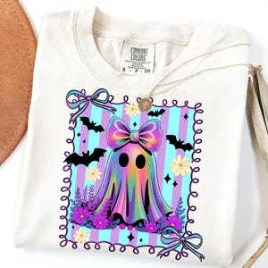 May include: Cream-colored t-shirt featuring a colorful ghost design with a large bow, bats, and flowers. The ghost has a rainbow gradient effect. The design is framed with a decorative border and bows. The shirt has the text "COMFORT COLORS".