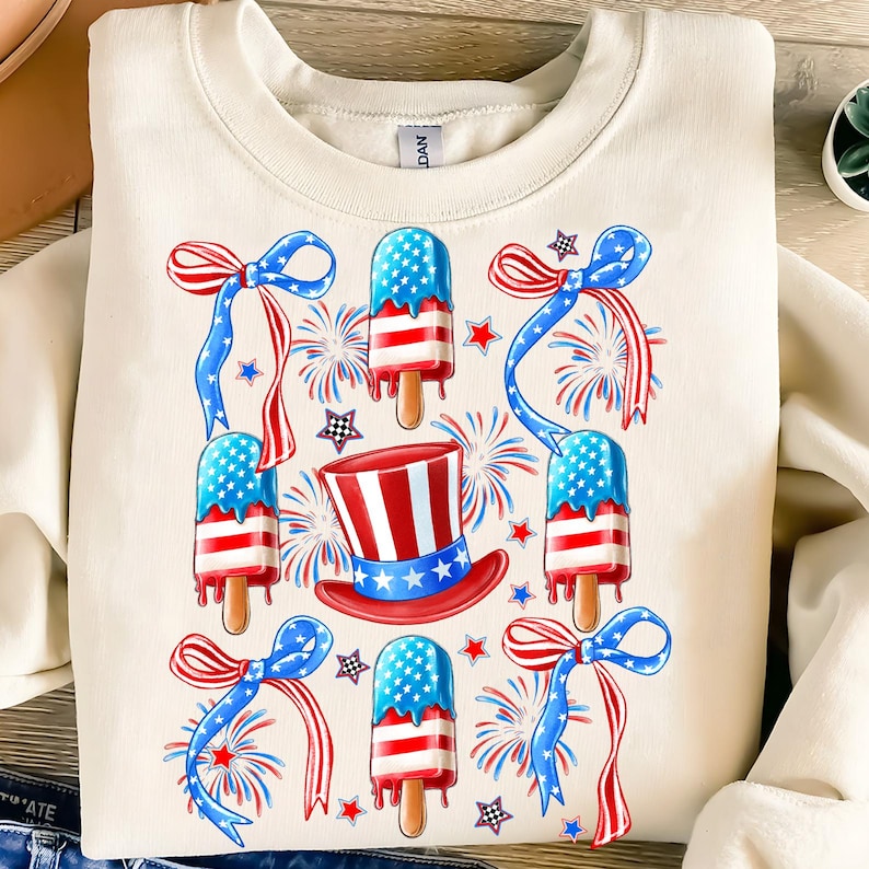 Puede incluir: Una sudadera de color crema con un dise&ntilde;o festivo que presenta paletas de hielo rojas, blancas y azules, un sombrero de copa y fuegos artificiales. Las paletas de hielo est&aacute;n decoradas con estrellas y rayas.