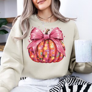 May include: Cream-colored sweatshirt featuring a colorful pumpkin design. The pumpkin is decorated with a mosaic pattern and a large pink bow. The design includes sparkling accents, perfect for fall or Halloween.