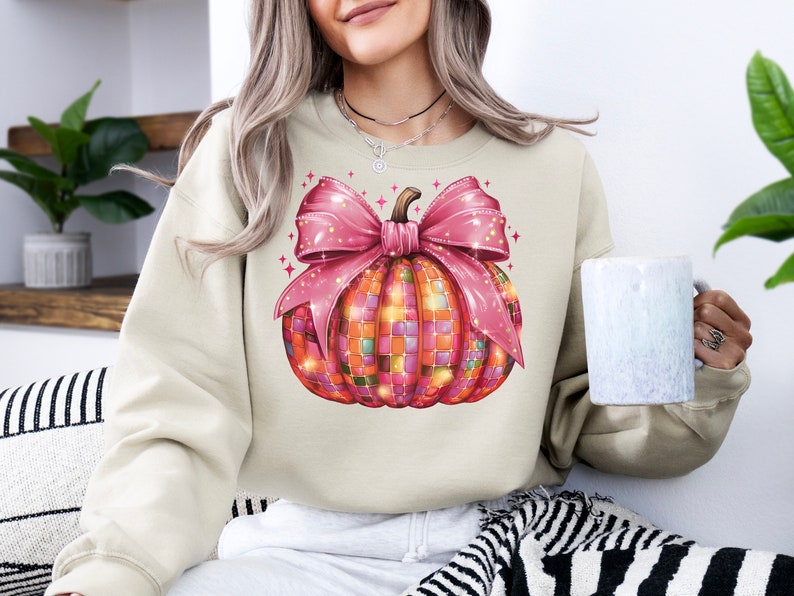 May include: Cream-colored sweatshirt featuring a colorful pumpkin design. The pumpkin is decorated with a mosaic pattern and a large pink bow. The design includes sparkling accents, perfect for fall or Halloween.