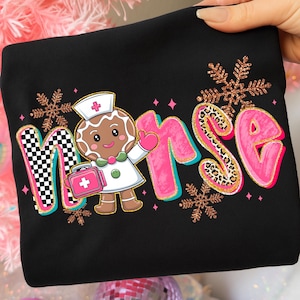 Pu&ograve; includere: Felpa nera con la scritta "Nurse" in un design giocoso. La "N" &egrave; a scacchi, la "urse" &egrave; rosa con accenti leopardati e presenta una grafica di infermiera di pan di zenzero. Fiocchi di neve glitterati dorati e diamanti rosa completano il design.