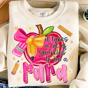 May include: Cream-colored sweatshirt with a colorful graphic. The graphic features a pink apple with a pencil, bow, and the text "it takes a lot of sparkles to be a para". Additional elements include stars, daisies, and a ruler.