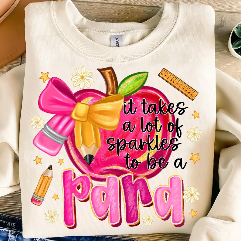 May include: Cream-colored sweatshirt with a colorful graphic. The graphic features a pink apple with a pencil, bow, and the text "it takes a lot of sparkles to be a para". Additional elements include stars, daisies, and a ruler.