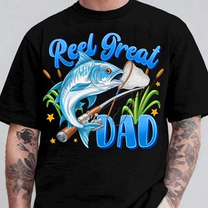 May include: Black t-shirt with a blue graphic of a fish, fishing rod, and the words "Reel Great Dad." The fish is light blue with orange accents. The text is in a stylized font with a blue gradient.