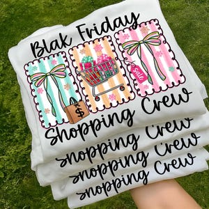 May include: White t-shirts stacked on top of each other, featuring the text "Blak Friday Shopping Crew." Each shirt has a colorful graphic with a shopping cart, gift boxes, bows, and a shopping bag with a dollar sign.