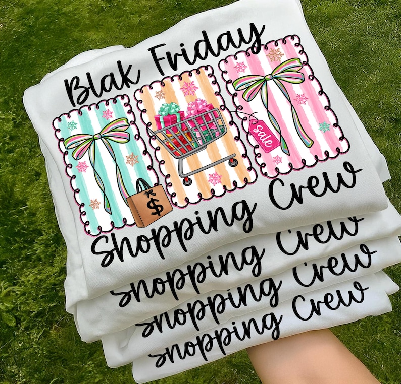 May include: White t-shirts stacked on top of each other, featuring the text "Blak Friday Shopping Crew." Each shirt has a colorful graphic with a shopping cart, gift boxes, bows, and a shopping bag with a dollar sign.