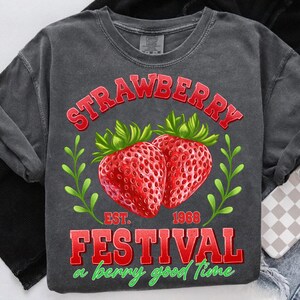 May include: A gray t-shirt with a red graphic design that says "Strawberry Festival" with the text "Est. 1968" and "a berry good time". The graphic also features two red strawberries with green leaves.