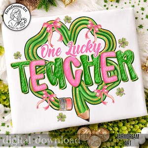 Może przedstawiać: Biała koszulka z zielonym wzorem koniczyny, złotymi brokatowymi akcentami i r&oacute;żowymi kokardkami. Napis "One Lucky Teacher" jest w kolorze r&oacute;żowym i zielonym. W projekt wkomponowano grafikę oł&oacute;wka. Na dole napis "digital download".