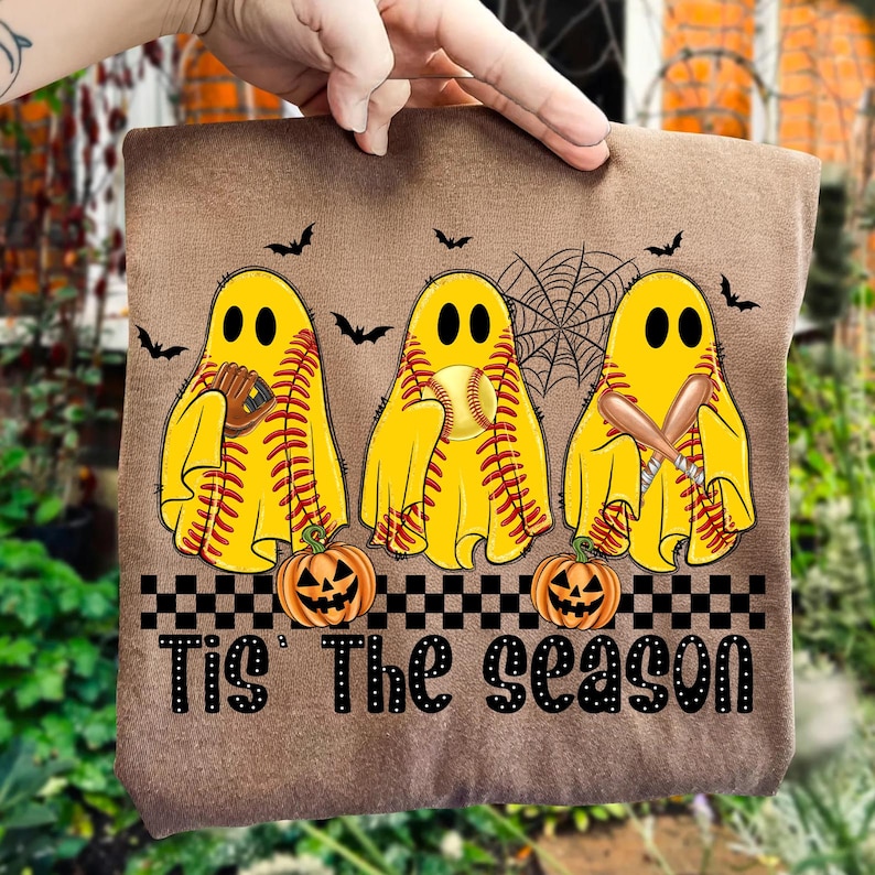 Puede incluir: Tela marr&oacute;n con un dise&ntilde;o de Halloween. Presenta tres figuras de fantasmas amarillos que sostienen equipos de b&eacute;isbol, bates y una pelota. Incluye murci&eacute;lagos negros, una telara&ntilde;a y calabazas. El texto "Tis The Season" est&aacute; impreso en la parte inferior.