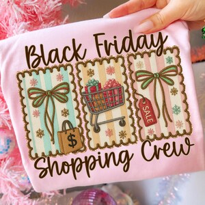May include: A light pink t-shirt with the words "Black Friday Shopping Crew" in brown script. The shirt features three decorative panels with bows, a shopping cart, a shopping bag, and a sale tag.