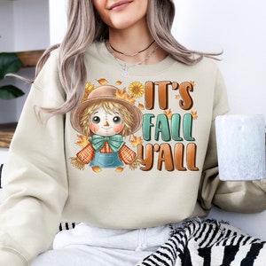 Op de afbeelding: Een beige sweatshirt met een cartoon-vogelverschrikker en de tekst "It's Fall Y'all".