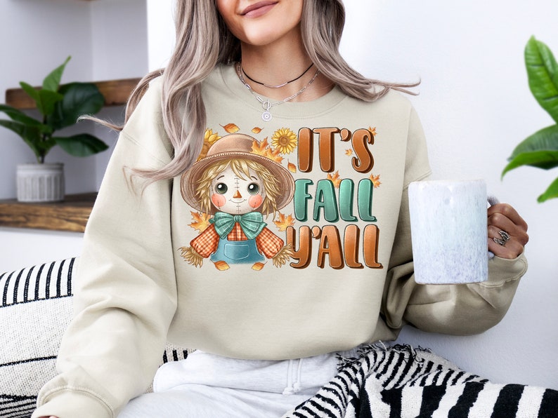 Op de afbeelding: Een beige sweatshirt met een cartoon-vogelverschrikker en de tekst "It's Fall Y'all".