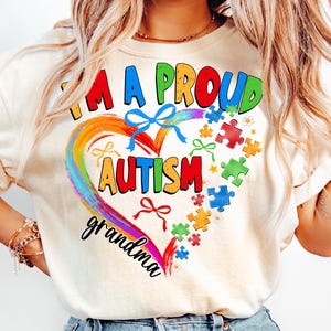 May include: A white t-shirt with a rainbow heart design and the text "I'm a proud autism grandma". The heart is surrounded by puzzle pieces and bows.