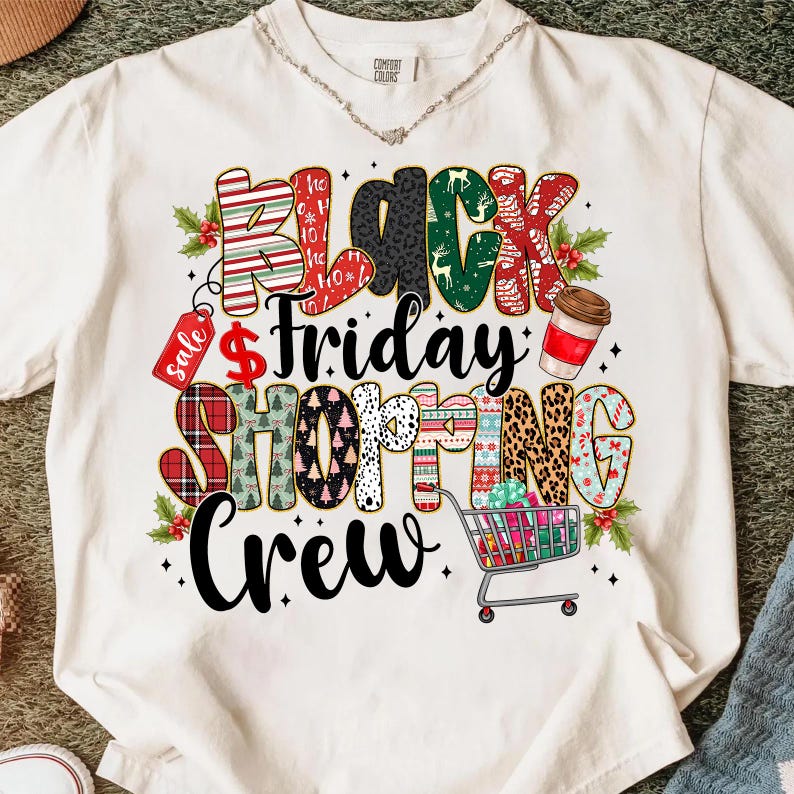 May include: Cream-colored t-shirt with a colorful graphic design. The design features the words "Black Friday Shopping Crew" in various patterns and colors, including red, green, and leopard print. A shopping cart and coffee cup are also included.