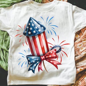 May include: White t-shirt featuring a watercolor illustration of a patriotic firework rocket. The rocket has a blue and white star-patterned cone, red and white striped body, and a red and blue bow. Blue and red fireworks are also depicted.