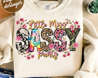 May include: A white sweatshirt with a design that says "Little Miss Sassy Pants". The letters are made of different patterns, including leopard print, camouflage, and tie-dye. The design is surrounded by pink flowers.