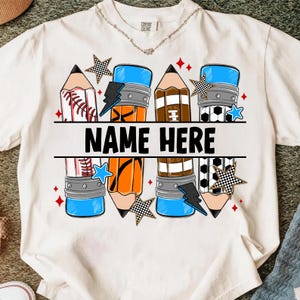 Pode incluir: Camiseta branca com um design de l&aacute;pis que incorpora elementos esportivos como beisebol, futebol americano e futebol. O texto "NAME HERE" est&aacute; em preto. Estrelas e raios decoram o design.
