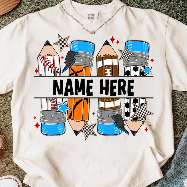 Pode incluir: Camiseta branca com um design de l&aacute;pis que incorpora elementos esportivos como beisebol, futebol americano e futebol. O texto "NAME HERE" est&aacute; em preto. Estrelas e raios decoram o design.