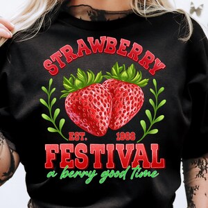 May include: Black t-shirt with a red and green graphic design featuring strawberries and the text "Strawberry Festival, a berry good time, Est. 1968".