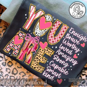May include: A dark gray t-shirt with the words "YOU ARE" in a decorative font, featuring pink hearts and patterns. The words "Enough, Brave, Worthy, Loved, Amazing, Strong, Capable, Smart, kind" are also displayed. The phrase "digital download" is at the bottom.