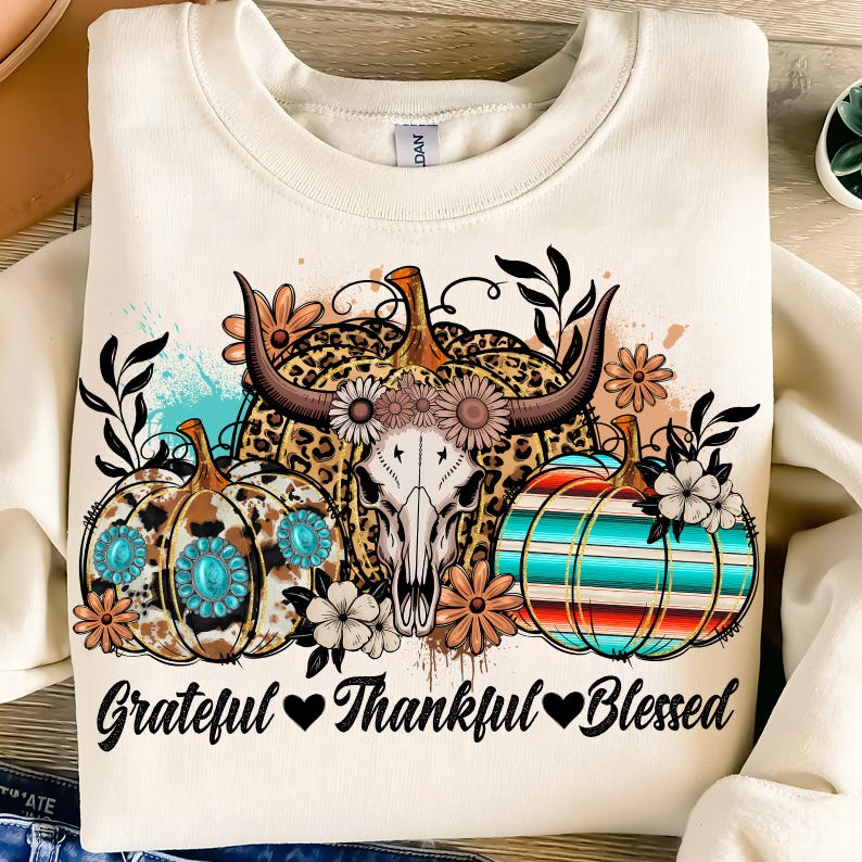 Puede incluir: Sudadera crema con un gr&aacute;fico tem&aacute;tico de oto&ntilde;o. El dise&ntilde;o incluye tres calabazas con varios patrones, una calavera de vaca y detalles florales. El texto "Grateful Thankful Blessed" est&aacute; impreso en la parte inferior.