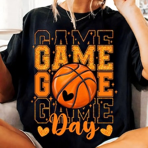 Peut inclure: T-shirt noir avec un texte en paillettes orange qui dit "Game Day" et un graphique de ballon de basket au centre.