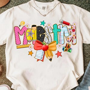 Può includere: T-shirt bianco con la scritta "Maestri" in lettere colorate e giocose. Il design include un righello, una matita, una mela e stelle, con dettagli di lampadina e fiori. La maglietta è del marchio Comfort Colors.
