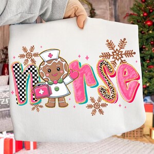 May include: White sweatshirt featuring the word "Nurse" in pink, teal, and leopard print, with a gingerbread nurse graphic. The design includes snowflakes and a checkered pattern, creating a festive, holiday-themed look.