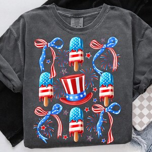 Puede incluir: Una camiseta gris oscuro con un dise&ntilde;o patri&oacute;tico que presenta paletas de hielo rojas, blancas y azules, un sombrero de copa y fuegos artificiales.