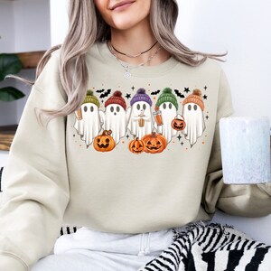 Op de afbeelding: Een beige sweatshirt met een afbeelding van vijf geesten die hoeden dragen en pompoenen en drankjes vasthouden. De geesten dragen hoeden in verschillende kleuren, waaronder paars, groen, oranje en bruin. Het sweatshirt is in een casual, comfortabele stijl.
