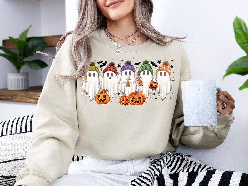 Op de afbeelding: Een beige sweatshirt met een afbeelding van vijf geesten die hoeden dragen en pompoenen en drankjes vasthouden. De geesten dragen hoeden in verschillende kleuren, waaronder paars, groen, oranje en bruin. Het sweatshirt is in een casual, comfortabele stijl.