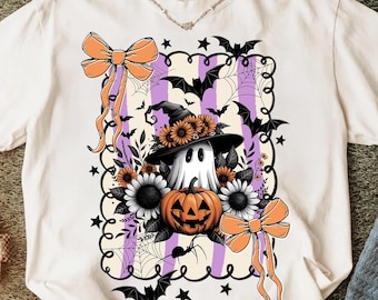 May include: Cream-colored t-shirt featuring a Halloween-themed graphic. The design includes a ghost wearing a witch hat, a jack-o'-lantern, sunflowers, bats, and bows. The graphic is set against a striped purple and white background.