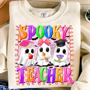 May include: Cream-colored sweatshirt with a colorful graphic that says "Spooky Teacher." The graphic features three cartoon ghosts, each holding school-related items like a book, pencil, and graduation cap. The design includes bats and spiderwebs.