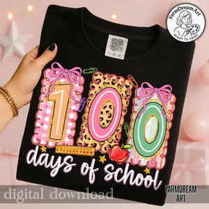 May include: Black sweatshirt featuring a colourful graphic celebrating 100 days of school. The design includes the numbers "100" in pink, orange, and green, with a ruler, apple, and the text "days of school".