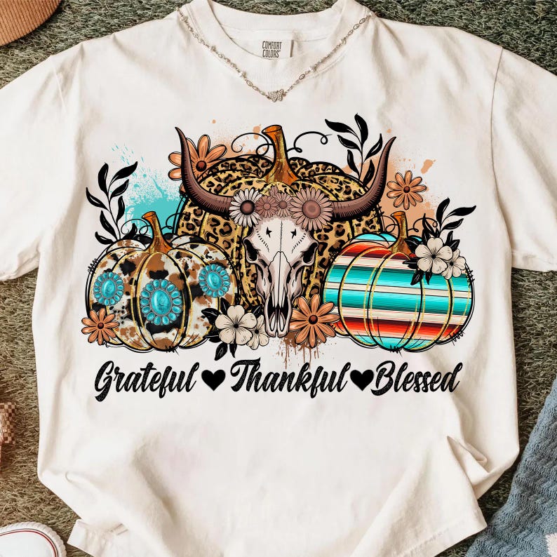 Puede incluir: Camiseta color crema con un gr&aacute;fico de tres calabazas con dise&ntilde;os florales y de animales, una calavera de vaca y el texto "Grateful Thankful Blessed". Las calabazas est&aacute;n decoradas con detalles turquesa y rodeadas de flores.