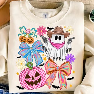 May include: Cream-coloured sweatshirt with a Halloween design. The graphic features a ghost in a cowboy hat holding guns, surrounded by pumpkins, bows, flowers, bats, and stars. The design is colourful and whimsical.