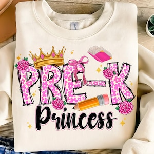 May include: Cream-colored sweatshirt with a graphic design for a preschooler. The design includes the words "PRE-K Princess" in pink floral print, a gold crown, pink roses, a bow, a book, and a pencil.