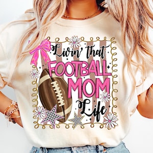 May include: Cream-colored t-shirt with a football-themed graphic. The design includes a football, pink bow, stars, and the text "Livin' that FOOTBALL MOM Life" in pink and black. The design is framed with a decorative border.
