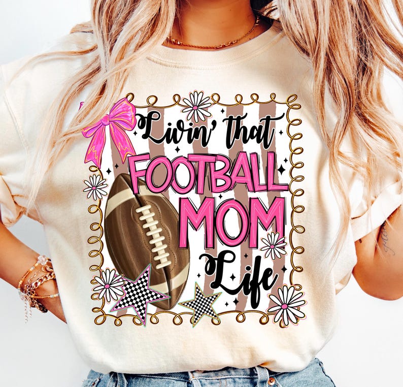 May include: Cream-colored t-shirt with a football-themed graphic. The design includes a football, pink bow, stars, and the text "Livin' that FOOTBALL MOM Life" in pink and black. The design is framed with a decorative border.