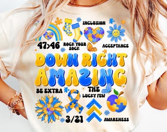 May include: A white t-shirt with a blue and yellow graphic design that says "Down Right Amazing". The graphic includes puzzle pieces, butterflies, sunflowers, and the text "47:46", "Rock Your Sock", "Inclusion", "Acceptance", "Be Extra", "The Lucky Few", "3/21", and "Awareness".