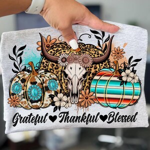 Puede incluir: Camiseta gris claro con un dise&ntilde;o gr&aacute;fico que presenta una calavera de vaca, calabazas y detalles florales. Las calabazas tienen patrones turquesas y a rayas. El texto "Grateful Thankful Blessed" est&aacute; impreso en la parte inferior.
