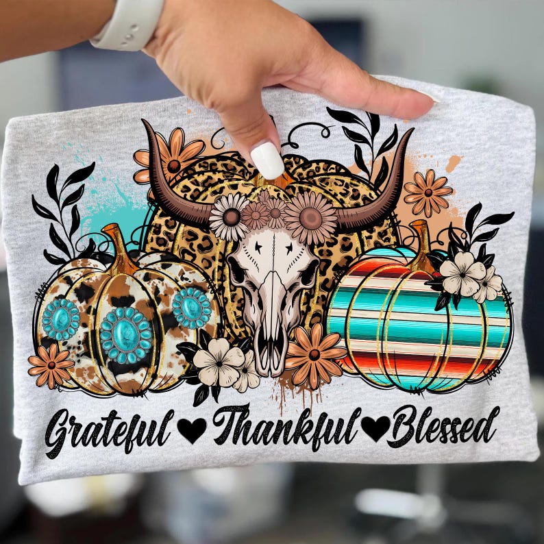 Puede incluir: Camiseta gris claro con un dise&ntilde;o gr&aacute;fico que presenta una calavera de vaca, calabazas y detalles florales. Las calabazas tienen patrones turquesas y a rayas. El texto "Grateful Thankful Blessed" est&aacute; impreso en la parte inferior.