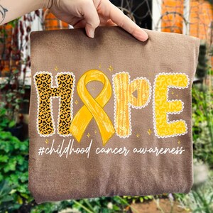 May include: A brown fabric item with the word "HOPE" in a mix of leopard print and yellow lettering, with a yellow ribbon in the center. The text "#childhood cancer awareness" is written below in white script.