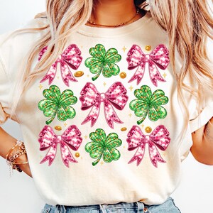 May include: A white t-shirt with a pink and green design. The design features four-leaf clovers and pink bows with gold accents.