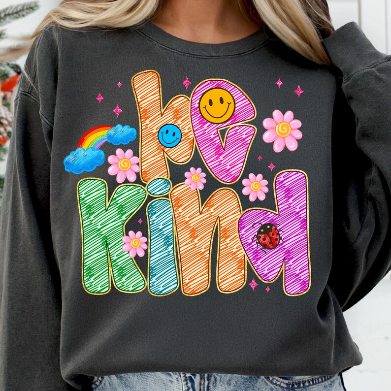May include: Dark gray sweatshirt with the words "be kind" in colorful, sketched letters. The design includes a rainbow, smiley faces, flowers, a ladybug, and star accents. The sweatshirt is a casual, comfortable style.