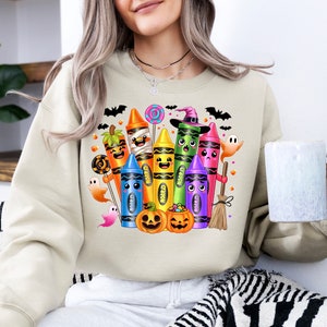 May include: Beige sweatshirt featuring a colorful Halloween-themed crayon design. The graphic includes various crayons dressed as Halloween characters, such as a mummy, witch, and pumpkin. Bats, ghosts, and candy complete the festive illustration.