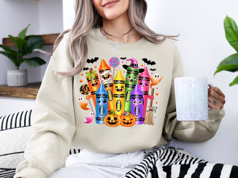 May include: Beige sweatshirt featuring a colorful Halloween-themed crayon design. The graphic includes various crayons dressed as Halloween characters, such as a mummy, witch, and pumpkin. Bats, ghosts, and candy complete the festive illustration.