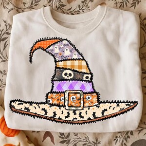 May include: White sweatshirt featuring a colorful patchwork witch hat design. The hat includes orange, purple, and black sections with skull, ghost, and floral patterns. The brim has a buckle with a skull and crossbones.